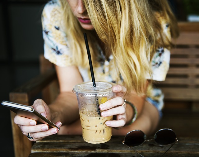 Een blond meisje die in een hand op haar telefoon zit en in de andere hand een beker ijskoffie vast heeft waar ze aan drinkt